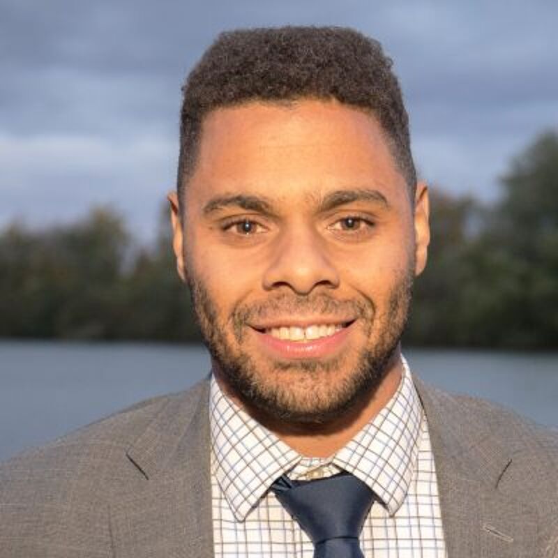 The image shows a close-up of a smiling man with a short haircut and a neatly trimmed beard. He is wearing a gray suit jacket, a checkered shirt, and a dark blue tie. The background is blurred, but it appears to be outdoors with a body of water and trees visible.